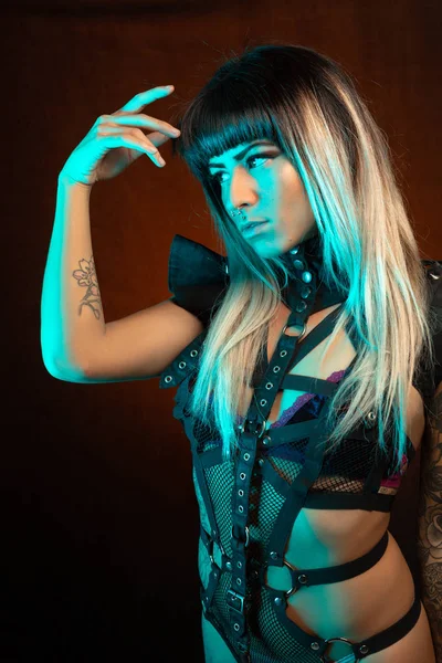 Alternative model with bangs and colored hair poses under blue light wearing a leather outfit