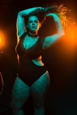 Renkli saç ve dantel bodysuit ve fileleri ile kıvrımlı alternatif model mavi ve turuncu aydınlatma altında pozlar