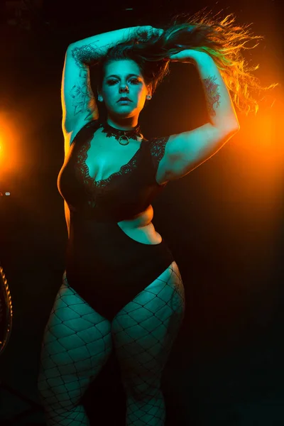 Curvy alternative model with colored hair and lace bodysuit and fishnets poses under blue and orange lighting