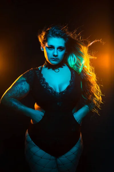 Close up portrait of curvy alternative model with colored hair and lace bodysuit and fishnets under blue and orange lighting