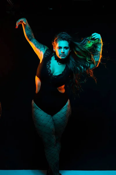 Curvy alternative model with colored hair and lace bodysuit and fishnets poses under blue and orange lighting