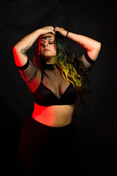 Curvy alternative model with colored hair and mesh clothing poses under red lighting