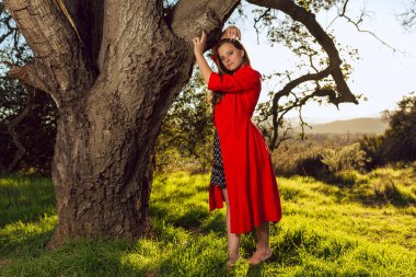 Kırmızı elbise giyen tan karışık ırk kadın üzerinde meşe ağacı günbatımında pozlar.