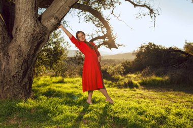 Kırmızı elbise giyen tan karışık ırk kadın üzerinde meşe ağacı günbatımında pozlar.