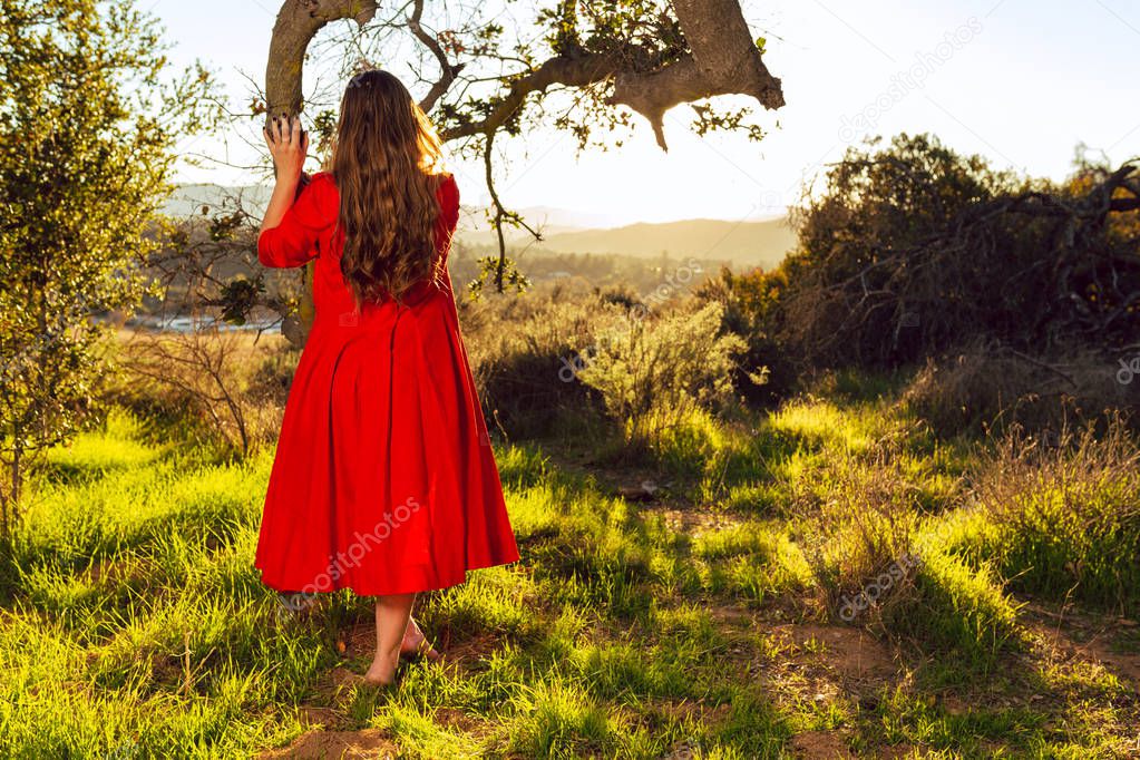 Tan mixed race woman wearing red dress poses on oak tree at sunset