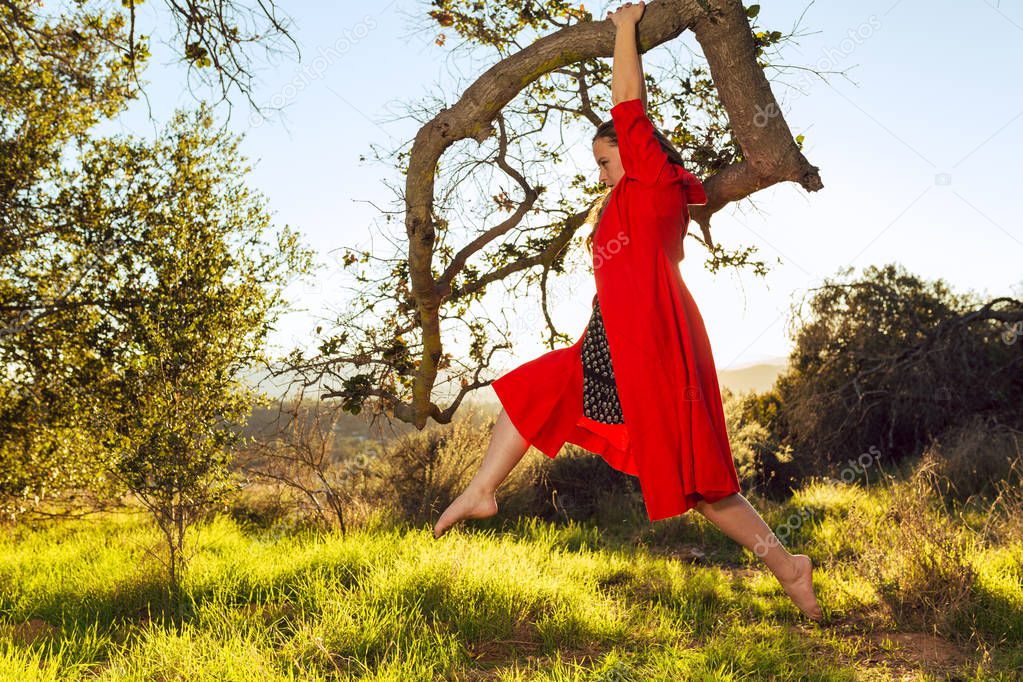 Tan mixed race woman wearing red dress does yoga hanging from oak tree