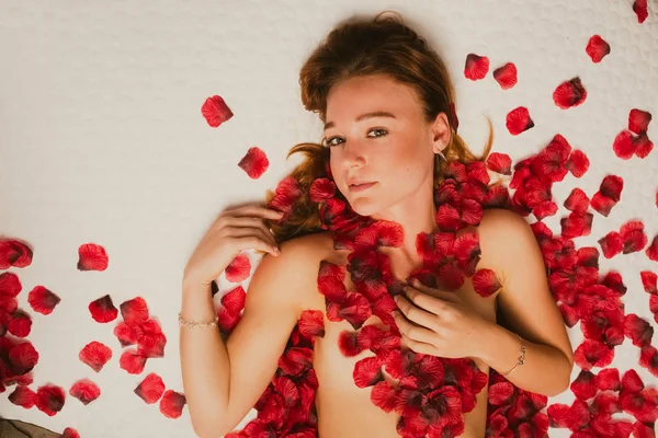 Redhead kaukasiska poserar på sängen täckt av rosenblad — Stockfoto