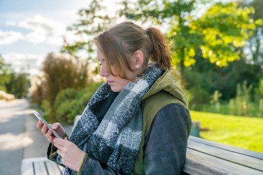 Genç bir kadın cep telefonu ile bir parkta bankta oturan ve kısa mesaj yazma