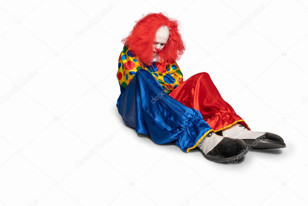 A sad clown is sitting on the floor looking down