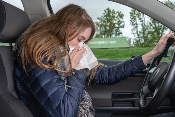 Una joven con alergia estornuda mientras conduce un coche en un pañuelo — Foto de Stock
