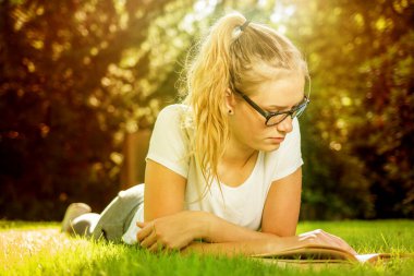 Bir kız çimenlerde uzanıyor ve kitap okuyor.