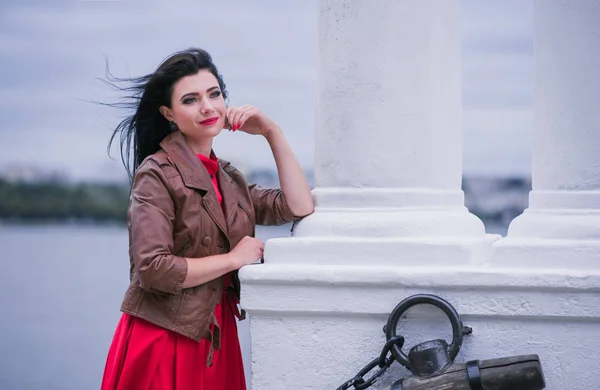 Gündelik kırmızı elbise ve göl şehir arka plan yürüyen kahverengi deri ceket genç moda kadın portresi. Güzel esmer model poz yatay portresi açık. Metin için yer