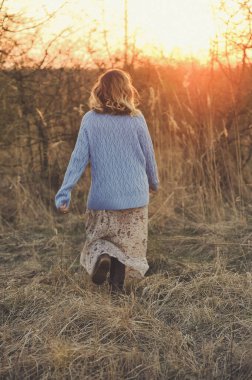 Mavi kazaklı ve çiçekli genç bir kadın gün batımında dışarıda yürüyor. Mutluluk, günbatımı arka planı