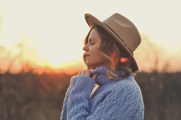 ベージュの帽子と青いセーターを着た若い女性の肖像画はリラックスしていて 日没時に暖かい公園で太陽を楽しんでいます 日没の背景 — ストック写真