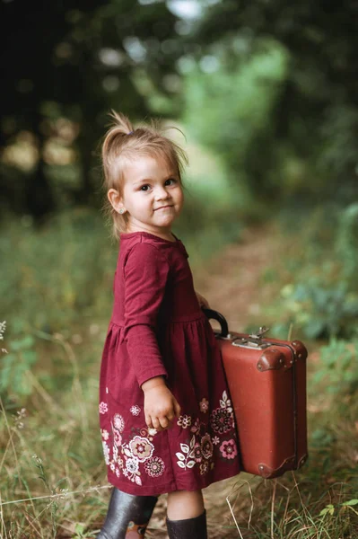 Burgundy Boho elbiseli güzel bir kızın portresi. Eski bavulunu yaz mevsiminde ormanda tutuyor. Retro tarzı. Doğa geçmişi, çocukluk, kartpostal