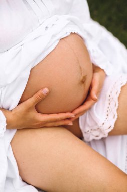 Hamile bir kadının karnının fotoğrafını çek. Beyaz yaz elbisesi içinde hamile bir kadın hamile karnını tutuyor. Aşk konsepti. Mutlu hamilelikler