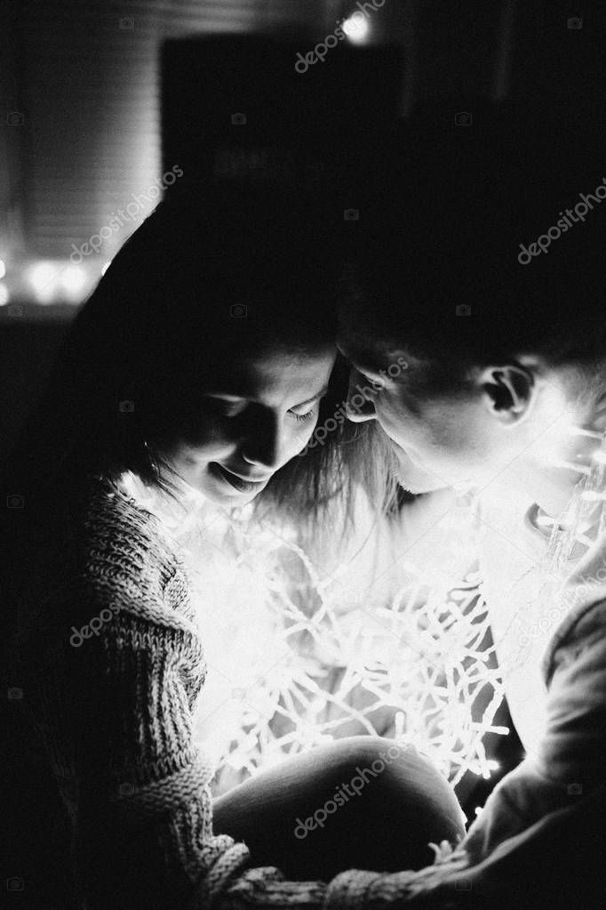 Loving couple hug each other on the bed wrapping in garland in bedroom. Black and white photo with noise filter