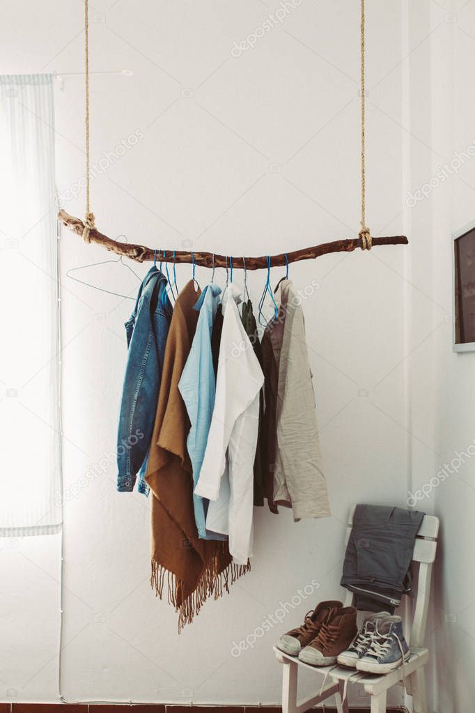 Many hangers with clothes on a wooden stick near shoes on chair