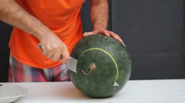 Turuncu gömlekli adam şef bıçağıyla karpuz kesiyor. Siyah arka plan