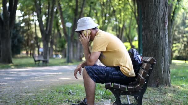戴帽子和太阳镜的人坐在公园的长椅上打电话说话 男子与电话交谈 而放松在树下树夏季 — 图库视频影像