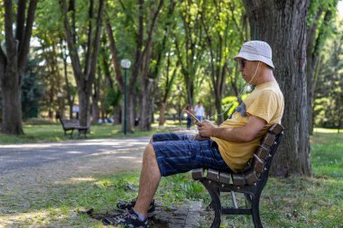 Şapkalı ve güneş gözlüklük adam parkta bankta otururken müzik dinler. Bir yaz günü ağaçların altında parkta dinlenirken adam telefonla müzik dinler