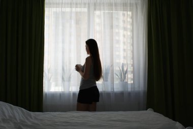 Bir kız elinde bir fincan kahveyle pencerenin yanında duruyor..