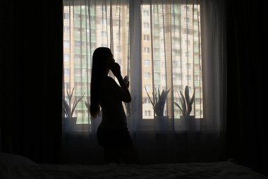 Pencerenin yanında akıllı telefonu olan bir kızın silueti..