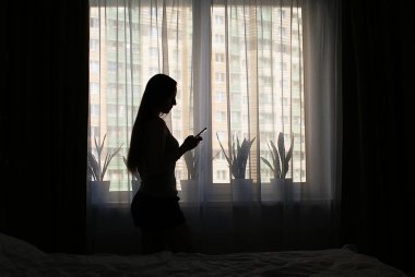Pencerenin yanında akıllı telefonu olan bir kızın silueti..