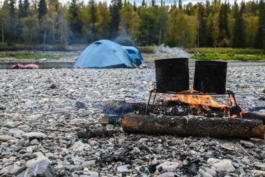 Kamp ateşi. Çadır ve teknelerin arka planında saksılar olan ateş.