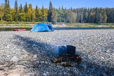 Kamp ateşi. Çadır ve teknelerin arka planında saksılar olan ateş.