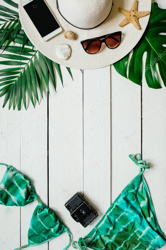 Bikini suit, hat, sunglasses, film camera, smartphone, sea star, green plam leaves arranged on wooden baclground. Summer holidays vacation concept. Vertical banner, postcard template.