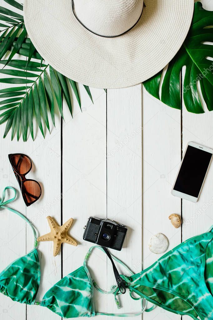 Bikini suit, hat, sunglasses, film camera, smartphone, sea star, green plam leaves arranged on wooden baclground. Summer holidays vacation concept. Vertical poster banner, postcard template.