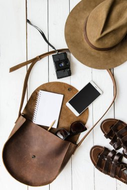 Kadın gazeteci veya seyahat aksesuarları Beyaz ahşap masa arka plan düz üzerinde yatıyordu. Kahverengi deri çanta, sandalet, kamera, cep telefonu, defter, kalem, ince tabaka fotoğraf makinesi. Metin için boş yer.