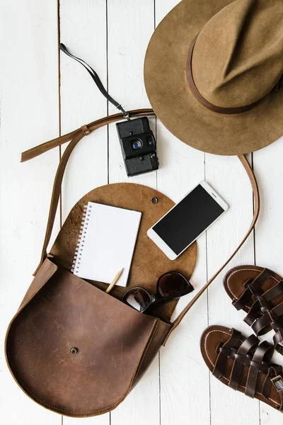 Kadın gazeteci veya seyahat aksesuarları Beyaz ahşap masa arka plan düz üzerinde yatıyordu. Kahverengi deri çanta, sandalet, kamera, cep telefonu, defter, kalem, ince tabaka fotoğraf makinesi. Metin için boş yer.