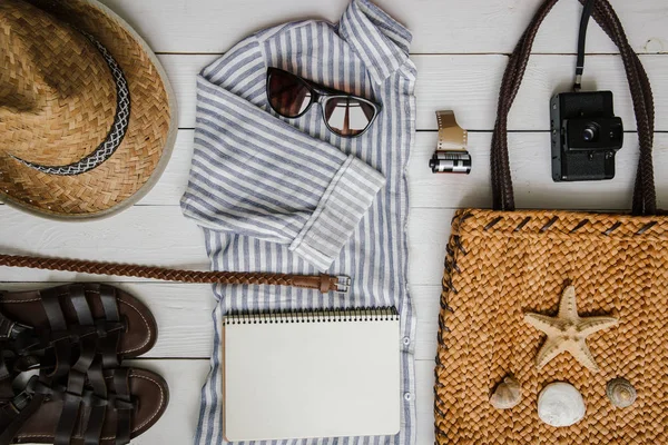 Kadının Akdeniz turistik giyim ve aksesuar beyaz ahşap masa arka plan düz üzerinde yatıyordu. Sarı organik saman Eko çanta, kahverengi deri sandalet, kemer, retro film kamerası. Boş defter alanı.