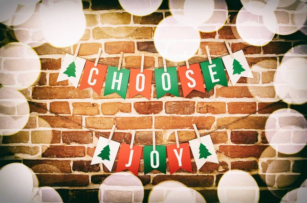 Red, green and white CHOOSE JOY paper card and rope homemade flag garland with top blurred lights border decoration on rustic yellow brown brick wall background. Nice vintage colored, hippster looking, horizontal greeting postcard.