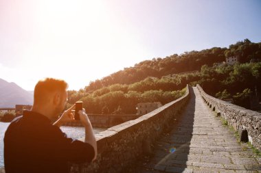 Adam bir Mozzano, Lucca, Toskana, İtalya karşısında nehrin Borgo Serchio Devils Bridge (Ponte della Maddalena) cep telefonunuzla resim alır. Doğal seyahat hedef kartpostal.