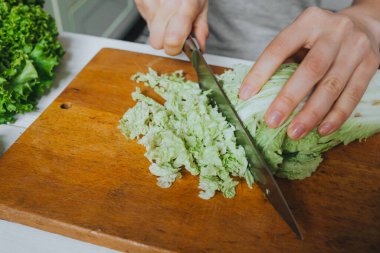Kadın elleri ahşap tahta üzerinde taze yeşil Pekin lahanası kesiyor. Beyaz masa arka planı. Keskin çelik bıçak yansıması..