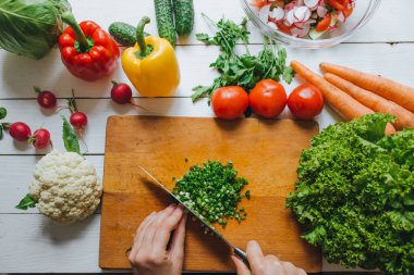 Kadın elleri tepeden bakıldığında tahta tahtada taze yeşil soğan kesiyor. Beyaz masa arka planı. Sağlıklı aşçılık kavramı. Etrafta sebzeler.