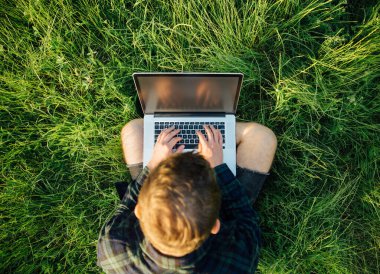 Genç hippi öğrenci gün batımında çimlerde dizüstü bilgisayar kullanıyor. Boş ekran kopyalama alanı. Üst genel görünüm.