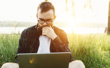 Modern sakallı, zeki, hippi bir erkek günbatımında çimlerin üzerinde oturuyor. Düşünceli bir şekilde dizüstü bilgisayara bakıyor.