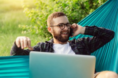 Neşeli, gülümseyen sakallı hippi turistler hamakta uzanmayı ve cep telefonuyla sohbet etmeyi severler. Teknoloji konsepti.