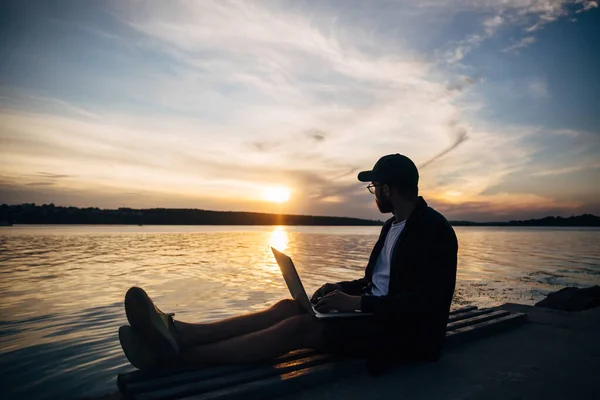 Silhouette of freelancer hipster male in baseball cap working on laptop on riverside in park and enjoying sunset. Room for text.