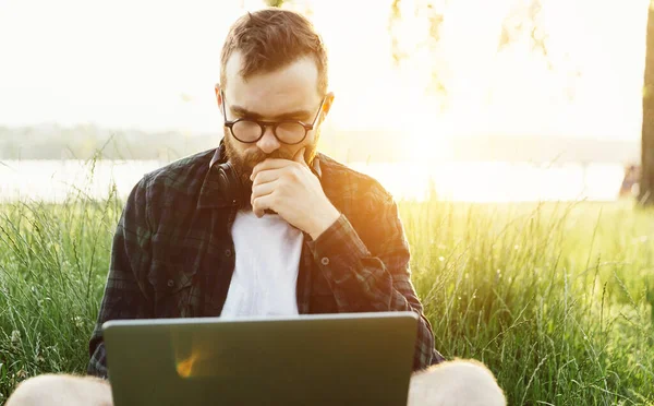 Modern sakallı, zeki, hippi bir erkek günbatımında çimlerin üzerinde oturuyor. Düşünceli bir şekilde dizüstü bilgisayara bakıyor.