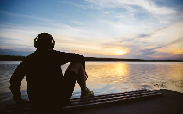Silhouette of man in headphones sitting on bench by the river. Enjoying music, online music streamin platform background concept.