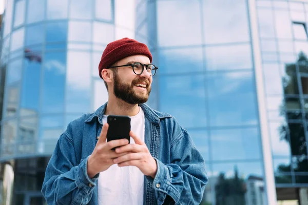 快乐而又英俊的年轻男性嬉皮士 拿着智能手机 朝旁边看去 微笑的男人随意的肖像 免版税图库照片
