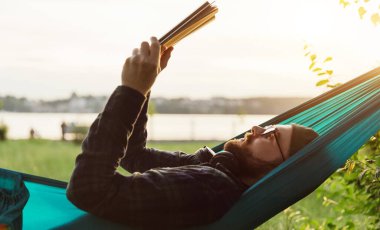 Sakallı hippi gözlüklü akıllı erkek gün batımında hamakta kitap okuyor. Boşluğu kopyala.