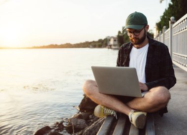 Günbatımında şehirdeki gölün kenarındaki bankta dizüstü bilgisayarda çalışan genç, sakallı, zeki, hippi bir adam. Boşluğu kopyala.