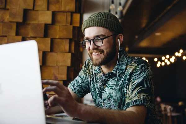 穿着眼镜和帽子 快乐而又成功的年轻帅气的嬉皮士男子坐在咖啡店里 带着笔记本电脑 面带微笑 图库图片