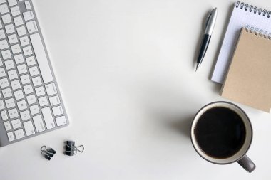 Beyaz ahşap masa klavye, kalem, defter, belge klipleri ve bir fincan kahve. Kopya alanı ile çalışma alanı üstten görünüm.